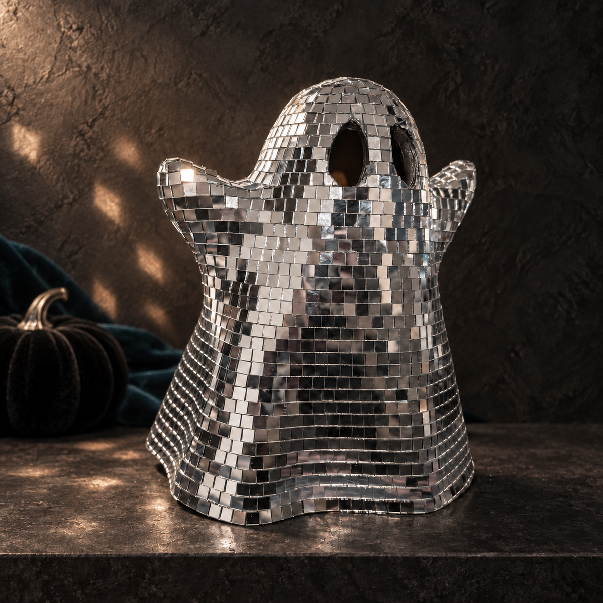 Silver glass mirror-tiled ghost without a bow on a dark stone console with petrol fabric and a black pumpkin.