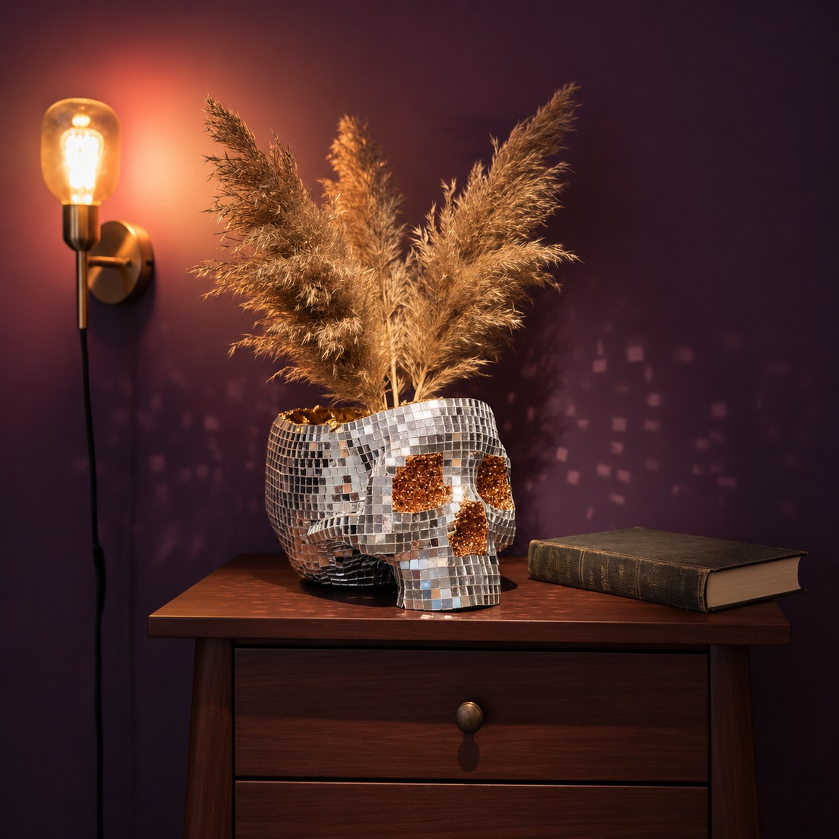 Disco skull planter with pampas grass on a dark bedside table beside a wall lamp and closed book