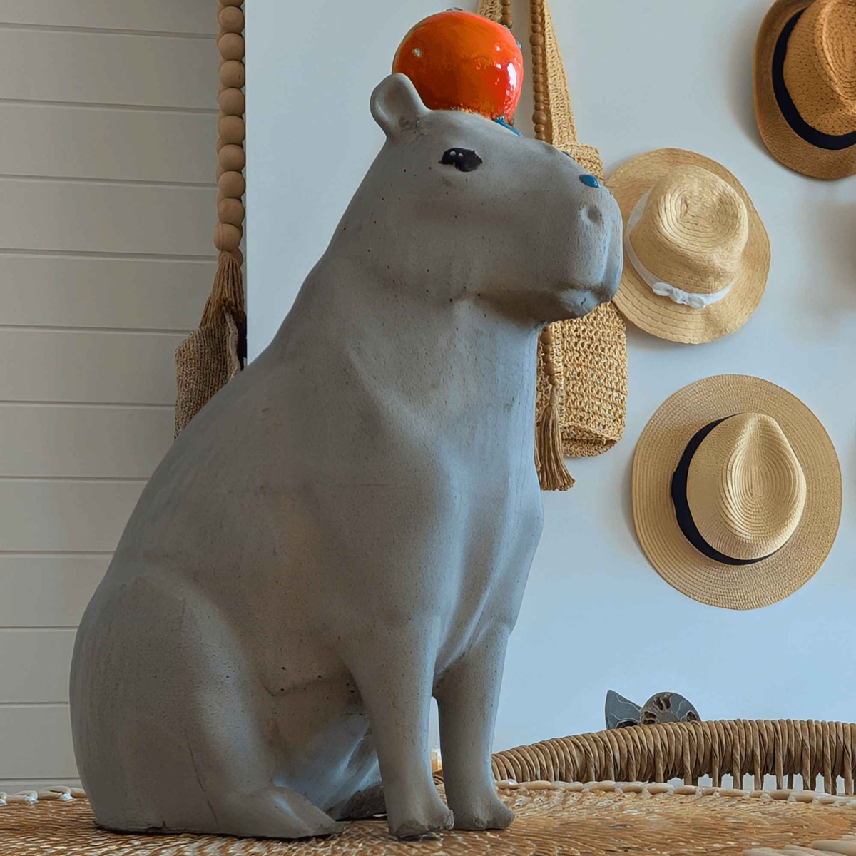 Cast stone capybara ornament displayed below a wall of straw hats